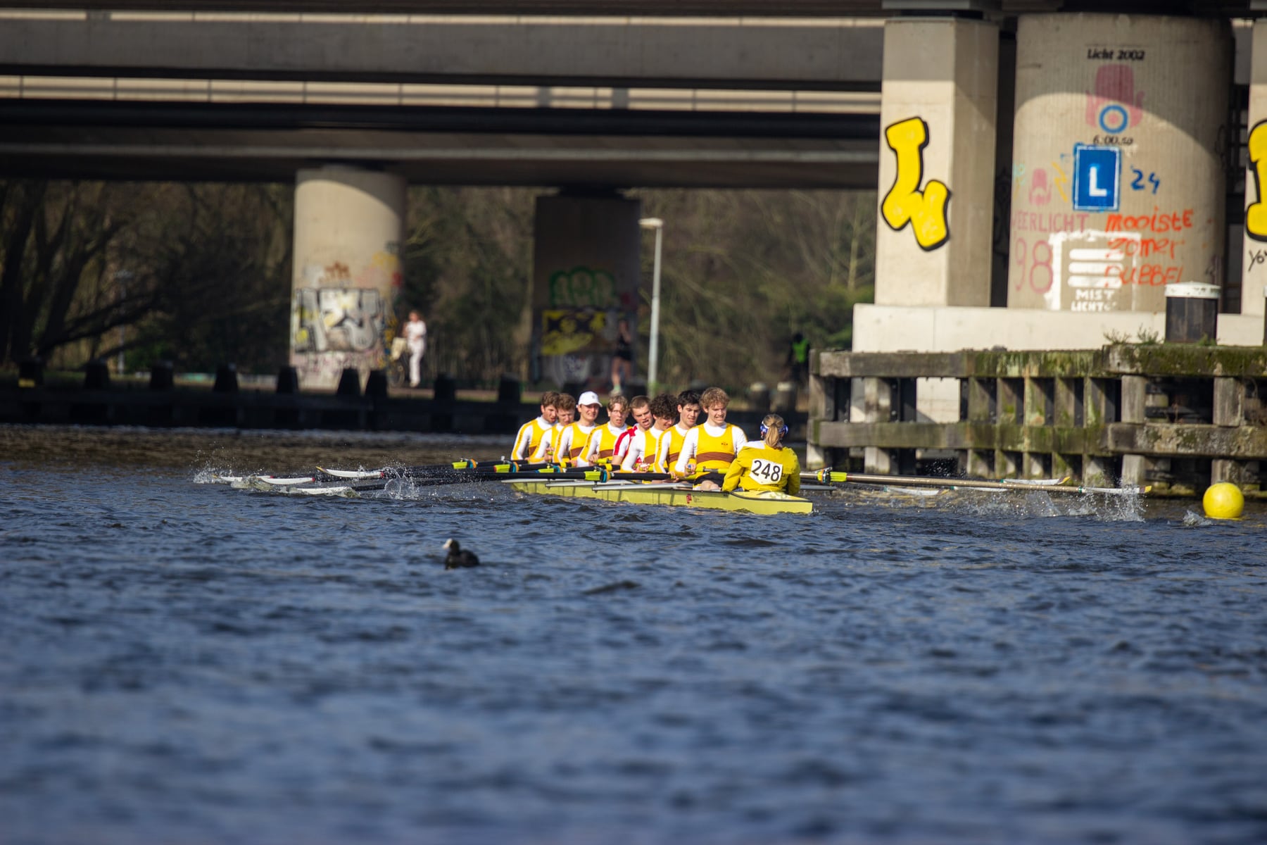 A distant crew rowing past a bridge with dark blue water in the foreground.