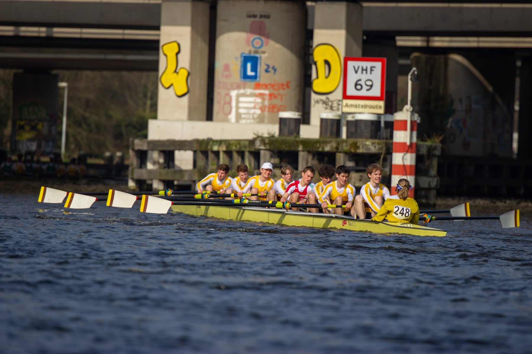 A yellow racing shell framed by bridge pillars and open water.