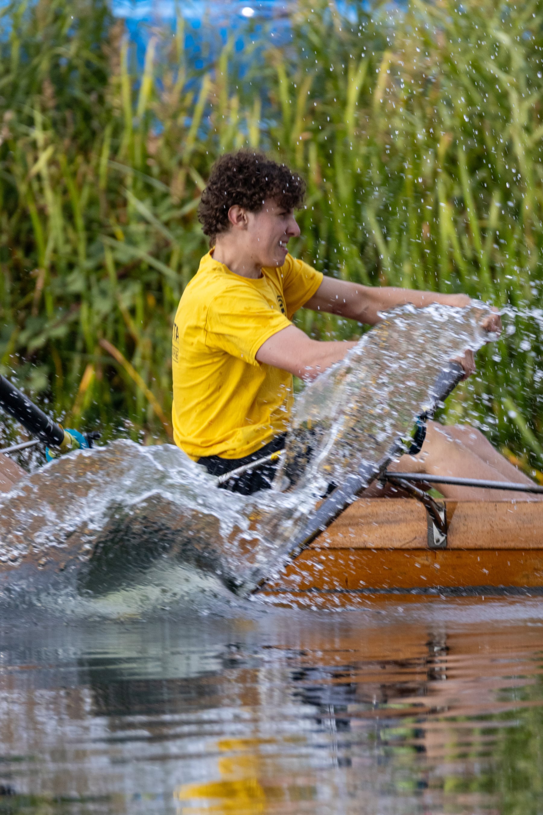 A rower throwing a sheet of water from a wooden shell in bright light.