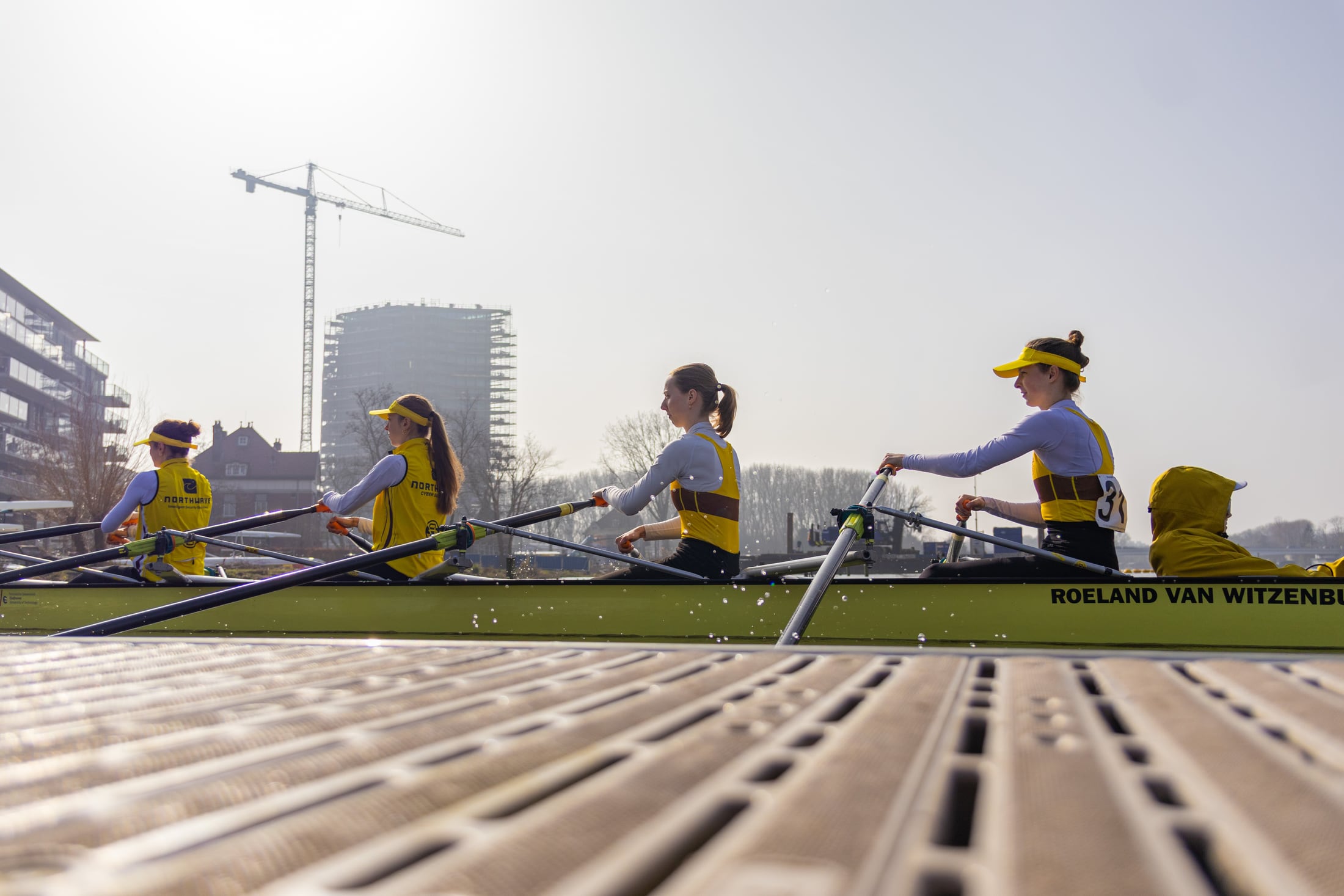 Low-angle rowing shell racing across dark Dutch water with warm light.